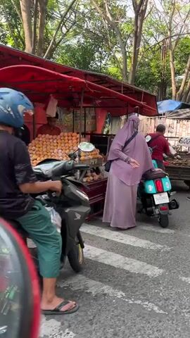 The video shows a bustling outdoor market scene. In the foreground, a woman in a purple hijab is standing next to a teal scooter. To her right, a man in a red shirt is attending to a vendor's stall, which displays various goods. Behind them, another man in a gray shirt is visible, and further back, more stalls and people are present. The stalls are covered with tarps and awnings. The environment appears to be a roadside market with trees and sunlight filtering through the leaves. The ground is paved, and there are markings on the road. The overall impression is one of lively commerce and activity.