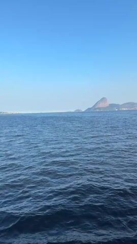 The video shows a wide shot of a calm blue ocean under a clear blue sky. In the distance, a large, rounded mountain rises from the water, with a city or town visible on its slopes. The water has gentle ripples and appears deep. The overall scene is serene and suggests a bright, sunny day.