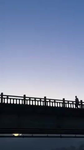 The video shows a vertical shot of the silhouette of a bridge against a gradient sky that fades from blue at the top to a lighter shade near the horizon.  Two individuals are seen walking on the bridge, their figures silhouetted against the sky. The bridge has a decorative railing with vertical supports. Below the bridge, there's a hint of something possibly reflecting light.