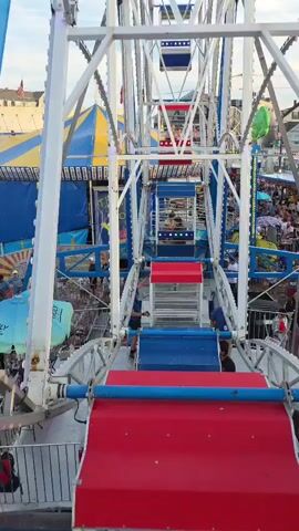 A vertical shot from inside a Ferris wheel shows the view of the amusement park as the wheel slowly ascends. Red and blue seats are visible in the foreground. People can be seen walking around below. In the background, a yellow and blue striped tent and other carnival attractions are visible.