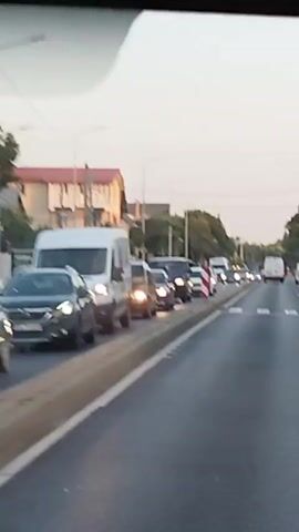 The video shows a traffic jam on a road. Cars and vans are stopped in multiple lanes. In the foreground, a white truck with a large trailer is stopped in the right lane. The truck has a red and white reflective marker on its rear bumper, and the word "SCHMITZ" is visible on the trailer. White crosswalk lines are painted on the road in front of the truck. In the background, there are more vehicles, streetlights, and trees lining the road. Buildings are visible on the left side of the road. The sky is a pale blue, suggesting it is daytime.