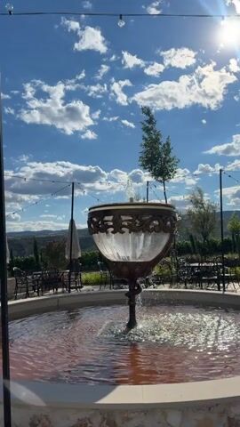 A video shows a fountain with water spraying up in the center, surrounded by a circular pool of reddish water. The fountain is ornately decorated with metalwork around a glass basin. In the background, tables and chairs are set up, suggesting an outdoor seating area for a restaurant or cafe. String lights hang overhead, and trees and hills are visible in the distance. The sky is bright blue with fluffy white clouds.