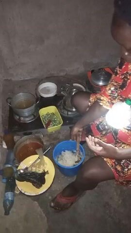 In what appears to be an indoor kitchen space, a woman wearing a short, red-and-black floral dress is seated on the floor preparing food. The walls are plain and the floor is a simple concrete material. She has a blue plastic bucket in her lap containing what appears to be mashed potatoes, which she is mixing with a wooden spoon. Several pots are arranged on a two-burner gas stove set on the floor. There is also a plastic container with chopped green vegetables and red peppers, and a plate with food, possibly fish and spaghetti, next to a pot of stew. A few plastic bottles are beside the stove. The lighting in the room seems dim and is coming from a hand-held source, casting a light on the bucket the woman is holding.