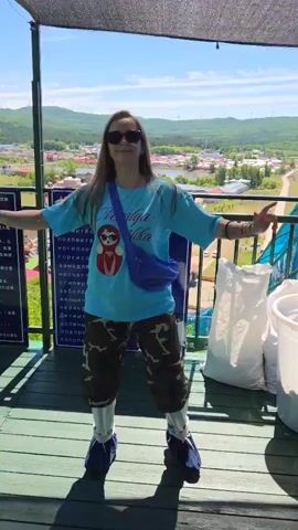A woman is featured dancing and posing on an elevated outdoor wooden platform during a bright, sunny day. Initially facing the camera, she then turns to face the scenic view behind her, continuing her dance. She is dressed in a light blue t-shirt with a graphic design that includes text like "Satiya" or similar, camouflage-patterned shorts, white leggings, and blue shoe covers over her footwear. A purple fanny pack is worn across her chest, and she has sunglasses on. Her long hair flows down her back as she moves.

The platform provides a panoramic view of a mountainous landscape with distant wind turbines under a clear blue sky. Below, a village with numerous houses and what appears to be a body of water is visible. A metal railing runs along the edge of the platform.

Behind the woman, a large informational sign is present, featuring text in Cyrillic script, which begins with "Меры предо..." (likely indicating "Precautions" or "Measures of precaution"). To the right of the woman, white sacks and white buckets are placed on the platform. The woman exhibits a joyful and playful demeanor as she moves, raising her arms and striking various poses. The strong shadows suggest midday sun. The location is contextualized as Jiejinkou, China.