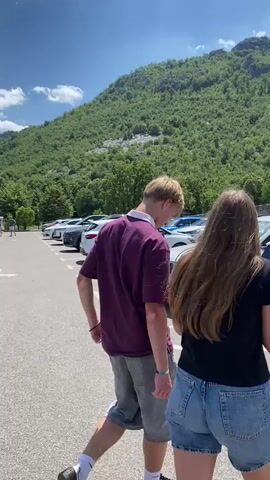 This video captures a sunny day in Lipa Dobrska, Montenegro, with a focus on people and vehicles in an outdoor setting. The scene appears to be a parking area adjacent to a lush, green mountainside.

Several cars are visible, parked in marked spaces, including white SUVs and sedans, suggesting a public or tourist destination. In the foreground, two young people, a male and a female, are walking away from the camera, with the male wearing a purple polo shirt and grey shorts, and the female in denim shorts and a black top.

Further in the background, other individuals and groups can be seen. Some are walking towards or away from buildings that suggest commercial establishments, possibly shops or restaurants, with a wrought-iron fence visible above one of them. A small, green trailer, possibly part of a tourist attraction or transport, is also present, with people gathered around it.

The weather is clear and bright, indicated by the strong sunlight and scattered white clouds against a deep blue sky. The presence of people dressed in summer attire, like shorts and light tops, reinforces the warm weather. The mountainous landscape, covered in dense green foliage, points to a natural or semi-rural environment.

A license plate is visible on the green trailer, reading "CT AR 595".

Overall, the video depicts a casual, leisurely scene with people enjoying a day outdoors in a scenic Montenegrin location. The atmosphere seems relaxed and pleasant, with a hint of activity around the parking area and the apparent tourist facilities.