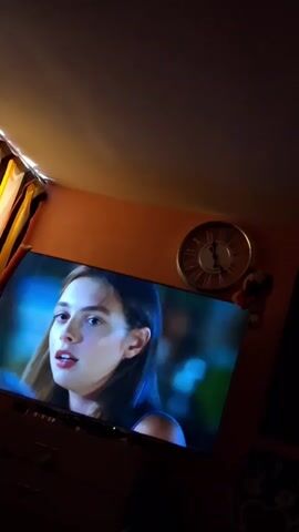 A television screen displays a close-up of a young woman's face with her mouth open as if speaking. She has long, dark hair and appears to be in a dimly lit room. To the upper right of the television, a round clock with Roman numerals is visible on an orange wall. A small teddy bear is perched on the wall next to the clock. Curtains hang on the left side of the frame, partially obscuring the scene. The overall lighting is dim and warm, emanating from the television and a light source above the curtains.