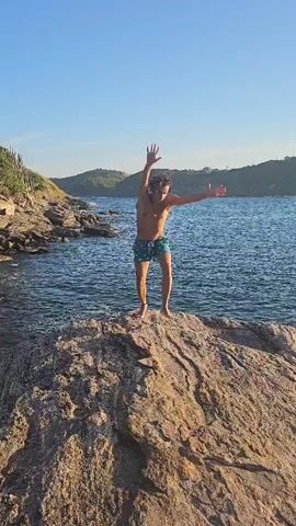 A young man does a handstand on a rocky outcropping by the sea. He then stands up and raises his arms in the air before walking out of frame. The scene is a sunny coastal environment with green hills in the background and blue water surrounding the rocks.