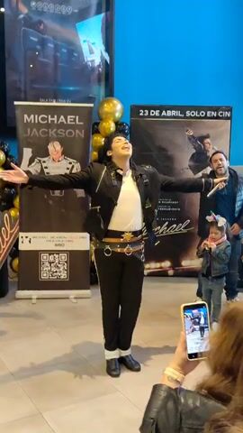 A person dressed as Michael Jackson stands on a tiled floor, striking a pose with arms outstretched, head tilted back, and mouth open as if singing. They are wearing a black jacket with embellishments, a white shirt, black pants with a decorative belt, and black shoes. Behind the performer, banners display images and text related to Michael Jackson, including "MICHAEL JACKSON" and "23 DE ABRIL, SOLO EN CINE". A string of gold and black balloons is also visible. To the right of the performer, a man holds a microphone, and a young child in a denim jacket looks on. In the foreground, a woman holds up a smartphone, appearing to record or photograph the scene. The environment appears to be an indoor public space, possibly a lobby or waiting area, with a blue wall in the background.
