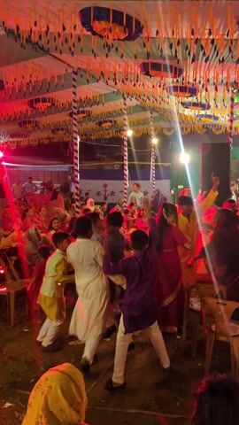 A group of people are dancing in a tent. There are men, women, and children dressed in colorful clothing, mostly pink, yellow, white, and purple. Some are seated on chairs around the dance floor. The tent is decorated with blue, green, orange and yellow streamers hanging from the ceiling. There are lights overhead, creating a bright atmosphere. A small stage is visible in the background. Some people are smiling and laughing. The overall atmosphere is festive and celebratory.