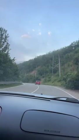 The video shows a view from inside a car driving on a winding road through a mountainous, wooded area. The road has two lanes with white dividing lines. There are two cars visible ahead, one white and one red, both moving in the same direction as the filming car. The sky is blue with some scattered clouds. Telephone poles are visible along the side of the road. The vegetation is dense and green, indicating a natural, possibly rural environment. The time of day appears to be late afternoon or early evening, given the lighting conditions.