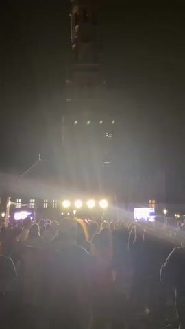 This is a vertical, low-angle nighttime video of a crowded outdoor concert in front of a large, historic building with a tall tower. In the foreground, a diverse crowd of people are gathered, facing a stage set up in front of the building. The stage is illuminated by bright lights, and a band or performers are visible on it. The building behind the stage appears to be a medieval or gothic-style structure, with a prominent clock tower reaching high into the dark sky. The atmosphere suggests a lively event, with people dancing and enjoying the music. The lighting shifts from blue to white, creating a dynamic visual effect.