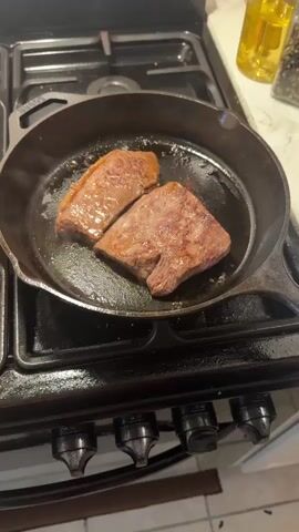 The video shows two pieces of steak being seared in a cast iron skillet on a gas stove. The steak is well-browned on the exterior, indicating it has been cooking for some time. The skillet is on a black gas stove with four burners and silver knobs. There is some oil or grease sizzling around the steak in the pan. In the background, a bottle of cooking oil and a jar of peppercorns are visible on a countertop. The lighting suggests an indoor kitchen environment. The video focuses on the cooking process of the steak.
