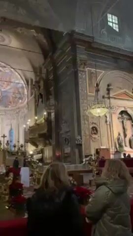The video shows the interior of a large, ornate church, likely in Europe, based on the architectural style. The camera pans around to show the high ceilings, a large dome with a painted fresco, and stained glass windows. Several people are seen walking around the church, including a priest near the altar. The church is decorated with poinsettias and golden ornaments, possibly for Christmas. The overall atmosphere is one of reverence and grandeur.