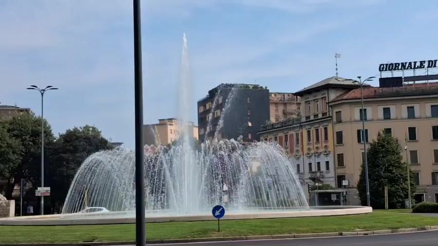 A large fountain sprays water in a circular pattern in a grassy median. A dark pole stands in the foreground. In the background, there are several buildings and trees. The sky is blue with some clouds. A white car is parked on the left side of the frame. A blue and white traffic sign is visible near the fountain. A street runs in front of the fountain.