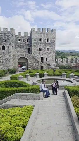 A man and two women are sitting on a bench in front of a stone building with crenellations. In the foreground, there are manicured bushes and a stone staircase. The scene is outdoors with a cloudy sky.