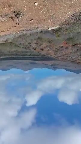 A drone's eye view of a dry, arid landscape with sparse vegetation and mountains in the distance. The sky is blue with white clouds. The camera is rotated 90 degrees counter-clockwise.