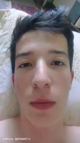 A close-up selfie video shows a young man's face. He has dark hair and is looking at the camera. His mouth is open and his tongue is sticking out. The background appears to be a bed with a patterned blanket. The timestamp indicates the video is very short, just a few seconds long.