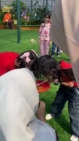 Two young girls are engaged in a game involving straws and rubber bands in a red bowl. One girl, with a pink bow in her hair, wears a red shirt and blue jeans. The other girl, with long dark hair, wears a red shirt and dark jeans. They are attempting to pick up rubber bands from the bowl using their straws. Several other children and adults are visible in the background on a green grassy area. Some children in the background are also participating in a similar activity, holding straws in their mouths. One adult, wearing a white hijab, is holding the red bowl steady for the girls.