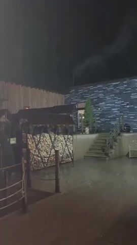 At night, the video shows a wooden outdoor deck in an outdoor establishment, likely a restaurant or bar. Stacks of wood can be seen in wooden shelters. A brick building is visible in the background. The camera moves to show a small outdoor bar with seating. The scene is dimly lit.