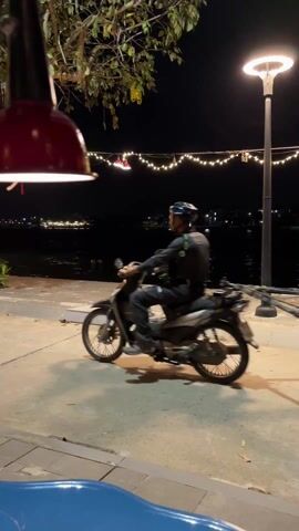 At night, on a paved path along a body of water, a man on a motorcycle pulls a trailer loaded with bicycles. The man wears a helmet and dark clothing. The trailer is blue and carries several bicycles with baskets. The background shows water with lights reflecting off it, and strings of decorative lights. A red, dome-shaped lamp hangs in the foreground, casting light on the scene. A blue object is partially visible in the lower left corner of the frame.