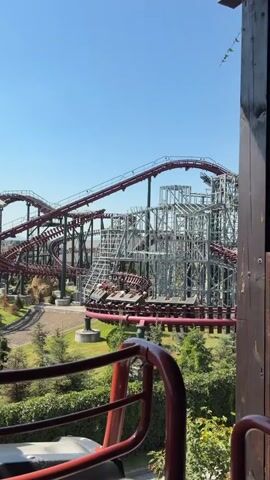 A video captures a roller coaster ride in an amusement park. The roller coaster track, primarily maroon and gray, curves through a landscape dotted with trees and some buildings in the background. A roller coaster train, carrying passengers, is visible moving along the track. The sky is clear and blue. The camera angle appears to be from another part of the roller coaster, looking out at the track and surrounding area. The first few seconds of the video show a wide shot of the roller coaster, followed by closer shots focusing on different sections of the track and a large, metallic structure integrated into the ride.