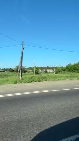 The video shows a view from a moving vehicle on a sunny day. The road has double white lines. On the left side of the road, there is a grassy area with some houses and trees in the background. A bus stop with a blue roof and yellow supports is visible. A woman in a light-colored dress is standing near the bus stop. Further down the road, there is a pedestrian crossing with yellow and white stripes. A brown metal fence runs along the right side of the road, and behind it are trees and some houses. The sky is clear and blue.