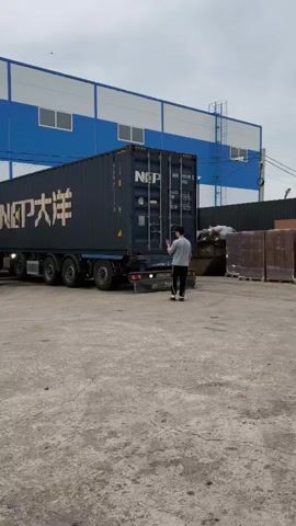 A man walks from the right side of the frame towards the back of a dark blue shipping container on a trailer. The shipping container has "NTP" and Chinese characters on it. He appears to be looking at the back of the trailer as he walks. Behind him, to the right, is a stack of boxed items. To the left is a large blue and white building with windows. The ground is paved. The sky is overcast.