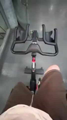 The video shows a person sitting on a stationary exercise bike. The camera is positioned above, looking down at the person's legs and the bike's handlebars. The person is wearing brown shorts and what appears to be a white t-shirt. They are actively pedaling. The bike has black handlebars with two water bottle holders. The floor is gray and has a checkered pattern.