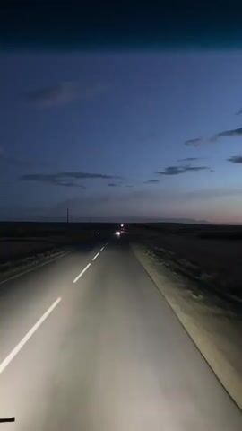 The video shows a first-person perspective of driving on a highway at dusk. The road is paved with a white line down the center and shoulders on either side. The sky is a deep blue with some clouds, indicating twilight or early night. The headlights of the vehicle illuminate the road ahead, casting bright beams. In the distance, other vehicles can be seen with their headlights on, and some faint red lights suggest tail lights. The surrounding landscape is dark and appears to be flat, with some vegetation and utility poles visible along the roadside. A thin wire or antenna is visible in the foreground, suggesting the viewpoint is from inside a vehicle, possibly a bus or truck. The overall mood is one of travel and transition, moving from daylight into darkness. The scene is set in a rural or semi-rural area, likely Baiqadam, Kazakhstan, given the provided location context. There are no people clearly visible, and no text is present in the video.