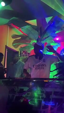 The video captures an energetic indoor party scene at night in Shenyang City, China. A DJ, wearing a white shirt with "Emirates FLY BETTER" printed on it and headphones, is actively mixing music at a DJ booth, with another person visible behind them. The venue features dim lighting with dynamic green and blue laser lights, creating a vibrant atmosphere. Large artificial green foliage adorns the walls and ceiling, contributing to a tropical or jungle aesthetic. Hanging lantern-style light fixtures are also visible. A prominent neon sign, reading "Oh My House" in a stylized script font, is mounted on a plant-covered wall, with green laser beams appearing to emanate from near it. Another partially visible sign reads "TIESTO RETERVILLE". People in the crowd are seen dancing and enjoying the music, indicating a lively nightclub or bar setting.