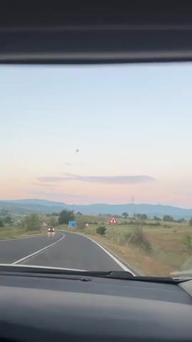 A view from the inside of a car driving on a road. The road curves to the left and goes uphill. There is a blue sign on the right side of the road, with white text and arrows. A single white car is driving in the opposite direction. The sky is a pale blue with pink and orange clouds, indicating it is either sunrise or sunset. Hills and trees are visible in the background. There is a triangular warning sign with a red border and a black symbol of a fallen tree on the right side of the road.