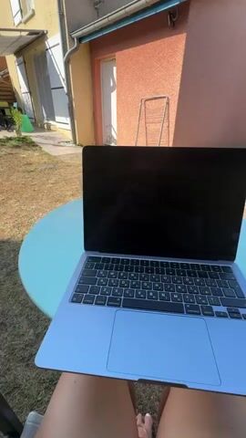 The video shows a person sitting in a backyard, with a laptop on a table in front of them. The backyard appears to be a residential setting with a house in the background. The grass is dry and yellow, suggesting it is a warm climate or season. The house is painted in warm colors, with windows and shutters visible. There is also a patio area with a table and chairs, covered by a pergola. The sky is clear and blue. The person's legs are visible in the foreground, wearing shorts. The laptop screen is black.