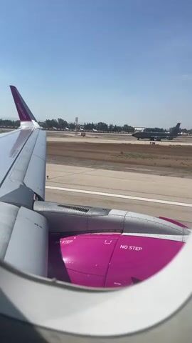 The video shows a view from an airplane window. The wing and engine of the plane are visible in the foreground. The engine is painted pink and has "NO STEP" written on it. The plane is on a runway at an airport, and another airplane is visible in the distance. The sky is clear and blue.