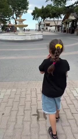 A girl with a yellow hair tie and a braid walks away from the camera on a paved walkway. She is wearing a black t-shirt and denim shorts. In the background, there is a large, multi-tiered fountain in the center of a circular plaza. Beyond the fountain, there are trees, buildings, and a body of water, likely the sea, under an overcast sky. People are visible walking around the fountain and in the distance. The scene appears to be a public outdoor space, possibly a promenade or park near the coast.