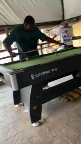 The video shows several young men playing pool on a Supermax Pro pool table in an outdoor setting, possibly a covered patio or veranda. One man in a green shirt is at the table, aiming with his cue. Other men are watching, some seated, some standing, and appear to be cheering or reacting to the game. The surroundings include a stone-tiled floor, a green and red striped rug, and various architectural elements suggesting a house or building. A white tent-like structure covers the area. The lighting is bright, and the atmosphere seems casual and jovial.