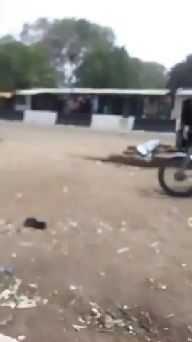 The video shows a man sitting on a motorcycle.  Another man gets off the motorcycle. The ground has some debris on it. There are trees in the background and a building. A man in a green shirt stands away from the motorcycle. Another man is seen holding a mobile phone, recording the scene.