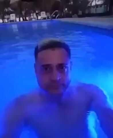 The video depicts a man waist-deep in a swimming pool. The pool water appears to be illuminated with a blue light. The man is looking directly at the camera, maintaining a relatively still position. In the background, there are trees and what appears to be lounge chairs. The scene is calm and suggests a nighttime or dimly lit setting.