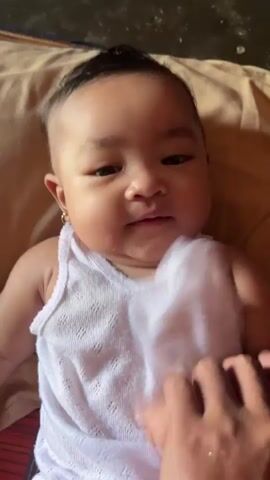 A close-up shot shows a baby lying on its back on a light brown blanket. The baby is wearing a white sleeveless undershirt with a textured pattern and white underwear with a red heart pattern. The baby has dark hair styled into a small mohawk and wears a small gold hoop earring in its left ear and a silver bracelet on its left wrist. Its right hand is raised and moving, and its left hand is resting on its chest. Two adult hands are visible at the bottom of the frame, one touching the baby's chest and the other near the baby's legs. The baby is looking directly at the camera with a neutral expression. The background is blurred, but appears to be indoors.