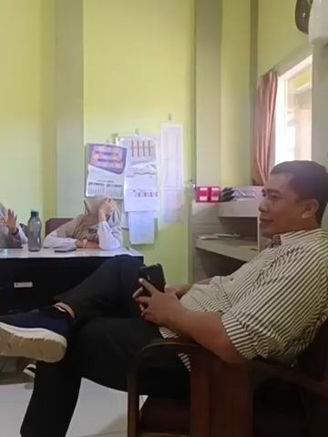 The video shows an indoor scene, likely an office or waiting area. Three women wearing headscarves are seated behind desks. The woman on the left is drinking from a red water bottle. The woman in the middle appears to be looking at something on the desk. The woman on the right is leaning forward with her hand on her head. In the foreground, a person's legs and feet are visible, suggesting they are seated in a chair. The room is painted light green, and the floor is tiled. There are various objects on the desks, including water bottles, papers, and a computer monitor. A framed document is hanging on the wall. The overall impression is one of a casual, possibly administrative, setting.