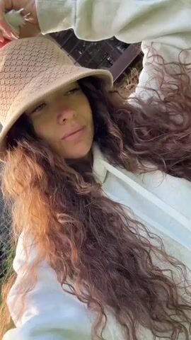 A woman with long, curly brown hair and a tan bucket hat is standing outside in a grassy area with trees in the background. She is wearing a white hooded sweatshirt and a tan crossbody bag with a patterned design. She is holding a white object, possibly a phone, in her right hand and appears to be recording a video of herself or taking a selfie. The background includes a building with a wooden exterior and a barbecue grill on a stone path. The lighting suggests it is daytime, possibly late afternoon or early evening, with sunlight filtering through the trees.