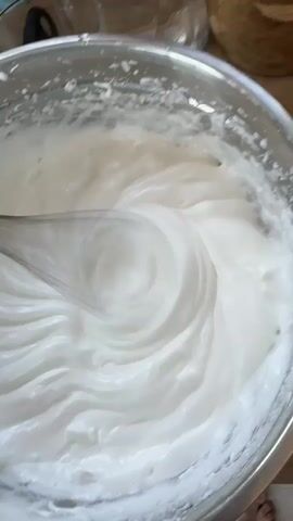 A close-up shot shows a stainless steel mixing bowl filled with a thick, white substance being whisked with a metal whisk. The substance appears smooth and glossy with distinct swirls from the whisking action. The whisk is moving steadily through the mixture. In the background are other kitchen items including glass bowls and a wooden bowl. The floor is visible, with a foot barely visible at the edge of the frame.