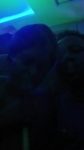 Two people are sitting close together in a dark room with overhead lights. The lights flash green and blue. One person is wearing a hat. Smoke is visible coming from a hookah. One person takes a drag.