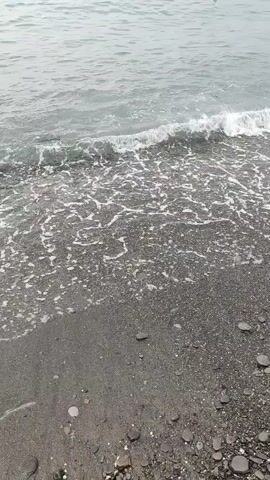 The video shows a view of waves gently washing ashore onto a dark, pebbly beach. The water is choppy and a light gray-blue color. The waves create white foam as they break on the shore. The beach is made up of small, dark stones and some sand. The overall scene is calm and natural, with no people or other objects visible. The camera appears to be held at a slight angle, looking down towards the water.