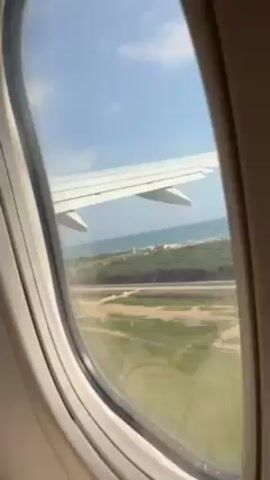 The view from an airplane window reveals an approaching coastline. The wing of a Ryanair plane is visible. The airplane is descending, with the coastline getting closer. A beach and a road parallel to the beach can be seen through the window. There are trees and vegetation on the land near the beach. The sea is visible in the distance.
