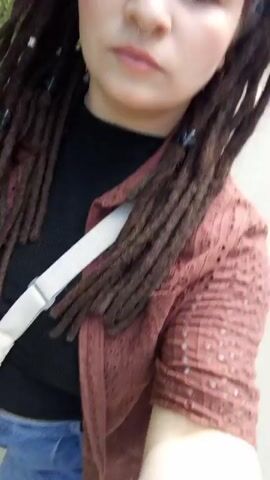 A medium shot captures a fair-skinned woman looking directly at the camera while walking on a paved path. She has brown eyes and a nose ring. Her dark hair is styled in dreadlocks, some of which are adorned with small, silver beads. A light grey headband with a dark pattern is wrapped around her forehead, pushing back some of her hair. She wears a black turtleneck shirt and a light brown, open-front cardigan with a lace-like pattern. A white strap of a bag or purse is slung across her body. The background consists of green foliage, suggesting a park or wooded area. The shot is slightly shaky, indicating it may have been taken while walking or moving. Other people are heard talking in the background.