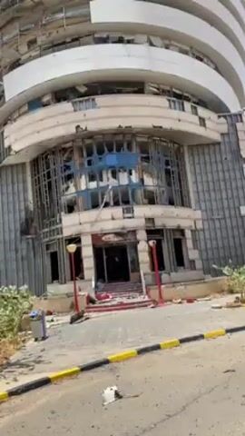 Alright, here's a concise description of the video:

The video shows a severely damaged high-rise building, seemingly from an explosion or structural failure. The building is a circular structure with multiple floors. Its facade is heavily damaged, with shattered windows and debris scattered around the entrance and surrounding area. The entrance area is particularly affected, with only the framework of the windows remaining. There's debris on the walkway leading to the entrance and on the street. The building's structure appears to be white with some blue and grey accents. There are red lampposts on either side of the entrance. A trash can can be seen near the walkway. The sky is visible and appears to be clear. The entire scene suggests significant destruction and damage.