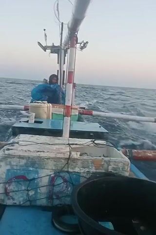 The video shows a person in a blue jacket sitting on a small fishing boat. The boat is on choppy water, with waves visible. In the foreground, there is a large black tub containing water and a fish. Next to the tub is a cooler box filled with fish, some of which appear to be small. The cooler box is white with blue and red markings. There is also a white plastic container on the boat. A person is seen in the background, huddled on the boat and appears to be wearing a blue jacket and hood. The sky is overcast.