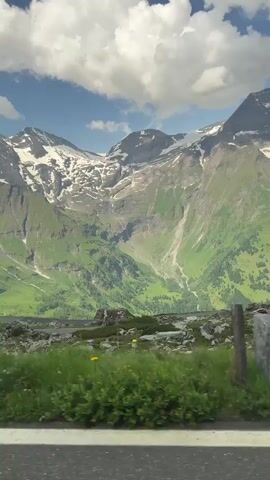 A first-person perspective video shows a cyclist on a bright yellow bicycle with a black rear derailleur, riding on a paved road. The cyclist is wearing a red shirt and black shorts. The background features a lush green mountainside with patches of snow on the peaks. The sky is blue with scattered white clouds. The video pans slightly to the right, showing more of the mountain range and the road ahead. A stone marker is visible on the side of the road.