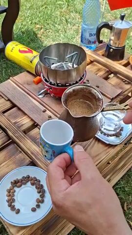 A person's hands are shown holding a small blue and white coffee cup with a patterned design and a blue handle. The person is pouring dark liquid from a metal pot, which appears to be a cezve, into the cup. The cezve contains a frothy, dark brown liquid, likely coffee. In the background, a portable camping stove with a silver metal windscreen is visible. Next to the stove is a can of yellow compressed gas fuel with red and black markings. To the right of the cezve is a small glass with a silver saucer, also containing a few coffee beans. In the foreground, to the left, is a white saucer with a pile of dark brown coffee beans. The scene appears to be outdoors, with green grass and wooden surfaces, suggesting a picnic or camping setting. A partially visible water bottle and a Moka pot are also in the background.