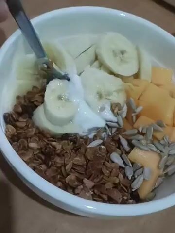 The video shows a close-up, slightly shaky view of a white bowl filled with a breakfast parfait. The bowl contains granola, sliced bananas, yogurt, cubed mango or cantaloupe, and sunflower seeds. A spoon is actively mixing the yogurt with some of the other ingredients. The lighting appears to be indoor, with a warm tone, and the background suggests a wooden surface, possibly a table. The focus is on the food within the bowl, with a small portion of a mug visible in the upper right corner, showing part of a red heart design.